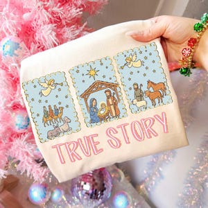 May include: Cream-colored t-shirt featuring a nativity scene illustration with angels, stars, and the text "TRUE STORY" in pink. The design includes images of the three wise men, Mary, Joseph, baby Jesus, and animals. The shirt has a festive, holiday theme.