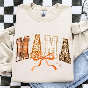 May include: Cream-colored sweatshirt with the word "MAMA" in a fall-themed design. The letters feature plaid, leaf, and leopard print patterns, with an orange bow and gold glitter accents. The sweatshirt is folded on a checkered surface.