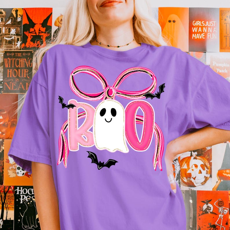 May include: A purple t-shirt featuring a Halloween-themed design. The design includes the word "BOO" in pink letters, a white ghost, black bats, and a pink and black bow. The shirt is a comfortable, casual style.