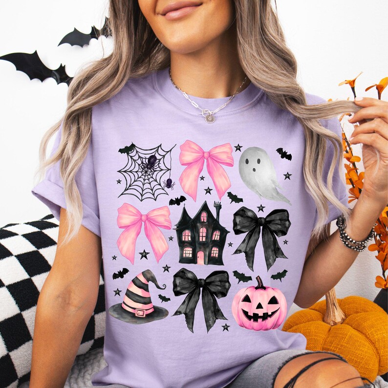 May include: A light purple t-shirt featuring a Halloween-themed design. The design includes a spider web, pink bows, a ghost, a haunted house, a witch hat, a black bow, a jack-o'-lantern, bats, and stars.