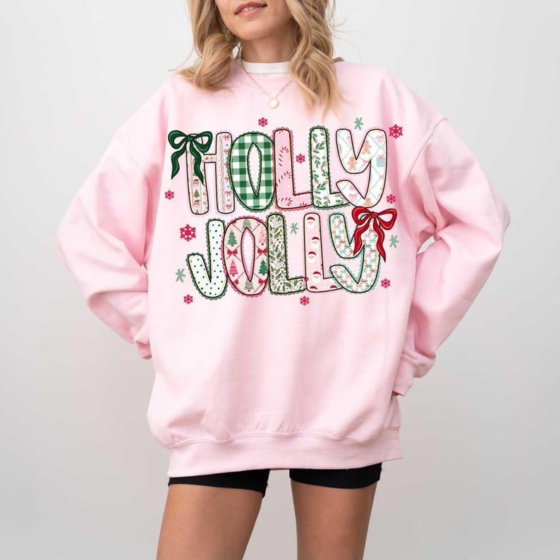 May include: A light pink sweatshirt with the words "HOLLY JOLLY" in a festive design. The letters are decorated with patterns, bows, and snowflakes. The sweatshirt is a long-sleeved crew neck.