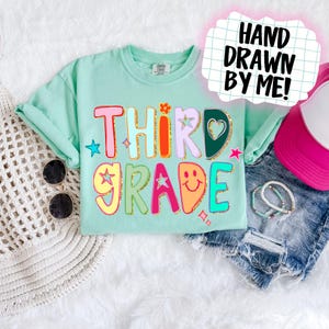 May include: A light teal t-shirt with the words "THIRD GRADE" in colourful, playful lettering. The design includes stars, a flower, and a heart. The text "HAND DRAWN BY ME!" is in a speech bubble. Sunglasses and a pink visor are also visible.