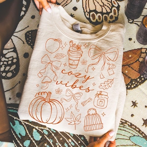May include: Cream-colored sweatshirt with a fall-themed design. The design includes a pumpkin, bows, leaves, a coffee cup, and the words "cozy vibes" in a handwritten font. The sweatshirt is held by two hands.