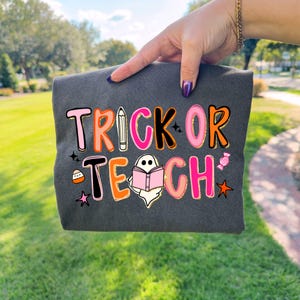 May include: Dark gray sweatshirt with the words "TRICK OR TEACH" in a colorful, playful font. The design includes a pencil, a ghost reading a book, and candy graphics, perfect for Halloween or teacher appreciation.