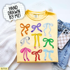 May include: A yellow t-shirt with a hand-drawn design of colorful bows. The bows are in various colors, including orange, purple, and green. The text "HAND DRAWN BY ME!" is in a speech bubble.