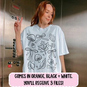 Op de afbeelding: Een lichtblauw t-shirt met een zwart-witte illustratie van paddenstoelen, strikken, bladeren en eikels. De tekst onderaan luidt: "COMES IN ORANGE, BLACK + WHITE. YOU'LL RECEIVE 3 FILES!"