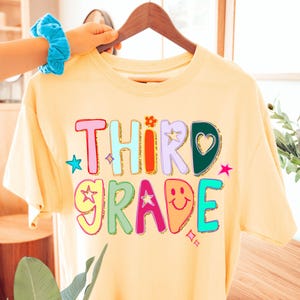 May include: A pale yellow t-shirt featuring the words "THIRD GRADE" in a vibrant, multi-coloured design. The design incorporates stars, a flower, and a heart. The t-shirt is displayed on a wooden hanger.
