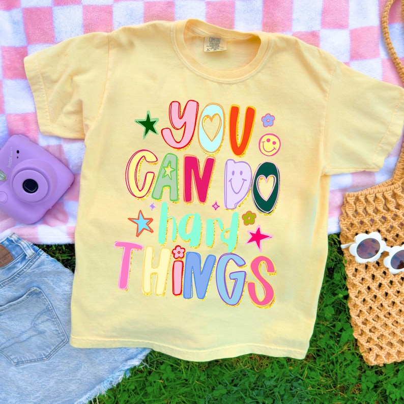 May include: A light yellow t-shirt with the words "YOU CAN DO HARD THINGS" in colorful, playful lettering. The design includes stars, hearts, and flowers. The shirt is on a pink and white checkered blanket with a purple camera and denim shorts.