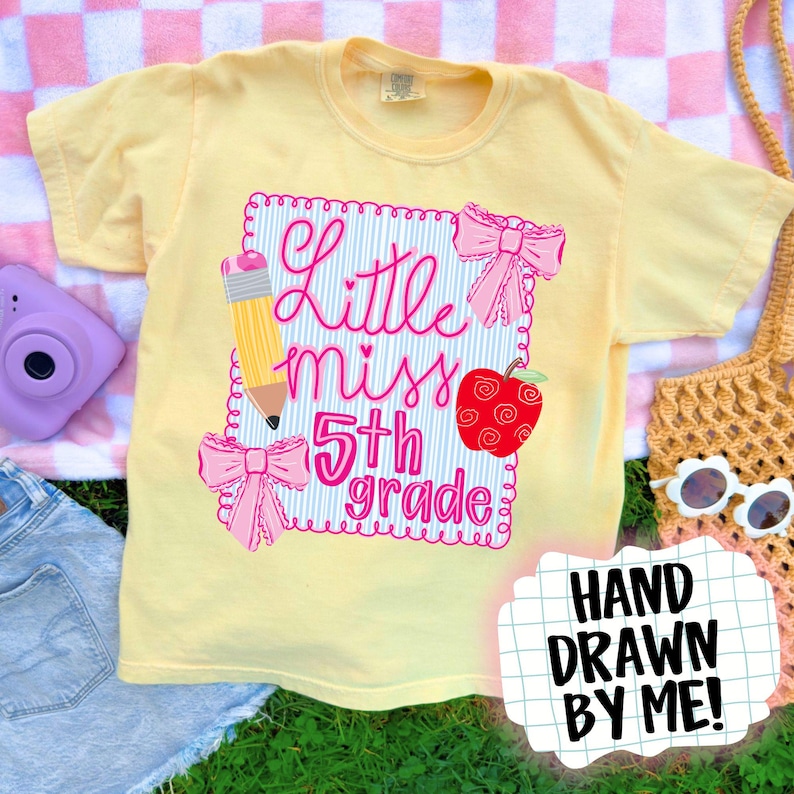 Op de afbeelding: Een lichtgeel T-shirt met een roze opdruk met de tekst "Little Miss 5th Grade". Het ontwerp bevat een potlood, een rode appel en roze strikken. De tekst "Hand Drawn By Me!" is ook zichtbaar.