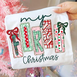 May include: White t-shirt with the text "my FIRST Christmas" in a festive design. The letters are decorated with patterns like polka dots, plaid, and candy canes, with bows on the "F" and "T".