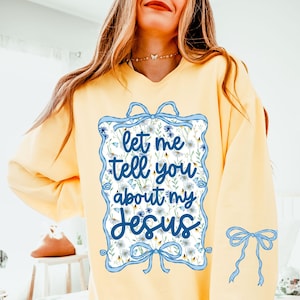 以下が含まれることがあります： 青い花柄のフレームと「let me tell you about my Jesus」というテキストが書かれた黄色のスウェットシャツ。フレームの上下には青いリボンが付いています。