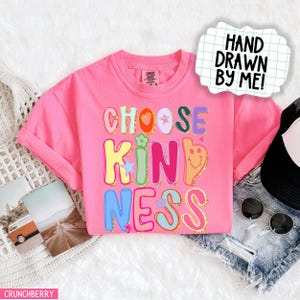 May include: A pink t-shirt with the words "CHOOSE KINDNESS" in colorful, bubble letters. The design includes a heart, star, and smiley face. The words "HAND DRAWN BY ME!" are in a white speech bubble.