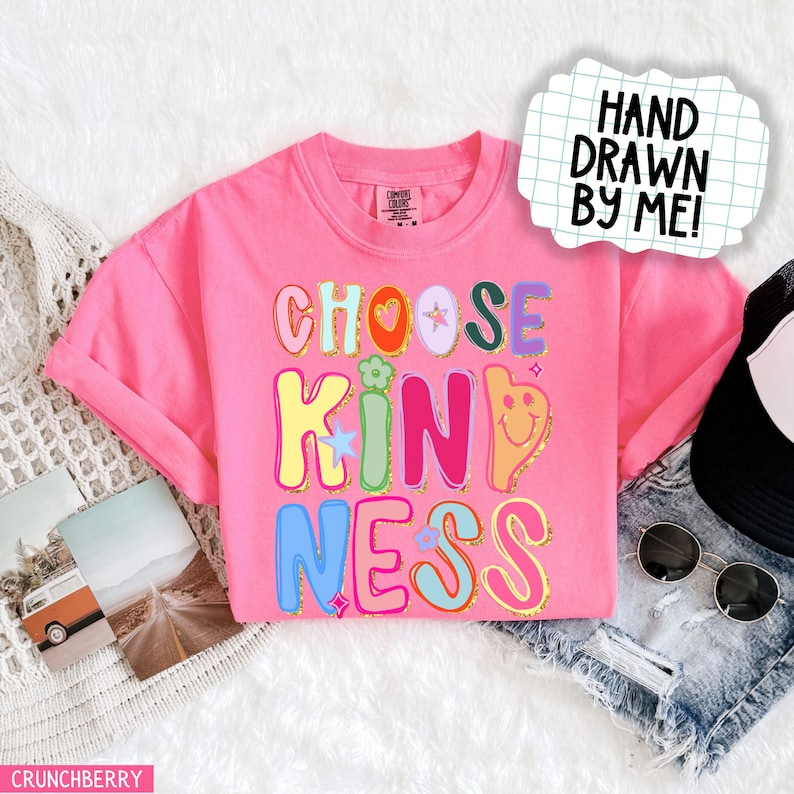 May include: A pink t-shirt with the words "CHOOSE KINDNESS" in colorful, bubble letters. The design includes a heart, star, and smiley face. The words "HAND DRAWN BY ME!" are in a white speech bubble.