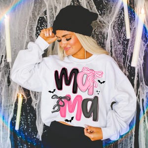 May include: White sweatshirt with the word "Mama" in black and pink, decorated with bats and bows. The sweatshirt is a long-sleeved crew neck. The person in the image is wearing a black beanie.