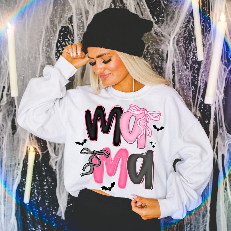 May include: White sweatshirt with the word "Mama" in black and pink, decorated with bats and bows. The sweatshirt is a long-sleeved crew neck. The person in the image is wearing a black beanie.
