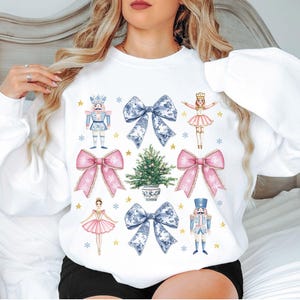May include: White sweatshirt featuring a festive design with illustrations of a nutcracker, bows, a Christmas tree, and ballerinas. The design includes pink and blue accents with gold stars and snowflakes, creating a holiday theme.