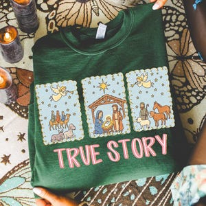 May include: A dark green sweatshirt featuring a three-panel design with a religious theme. Each panel has a light blue background with a gold scalloped border and illustrations of angels, stars, and biblical scenes. The words "TRUE STORY" are printed in pink.