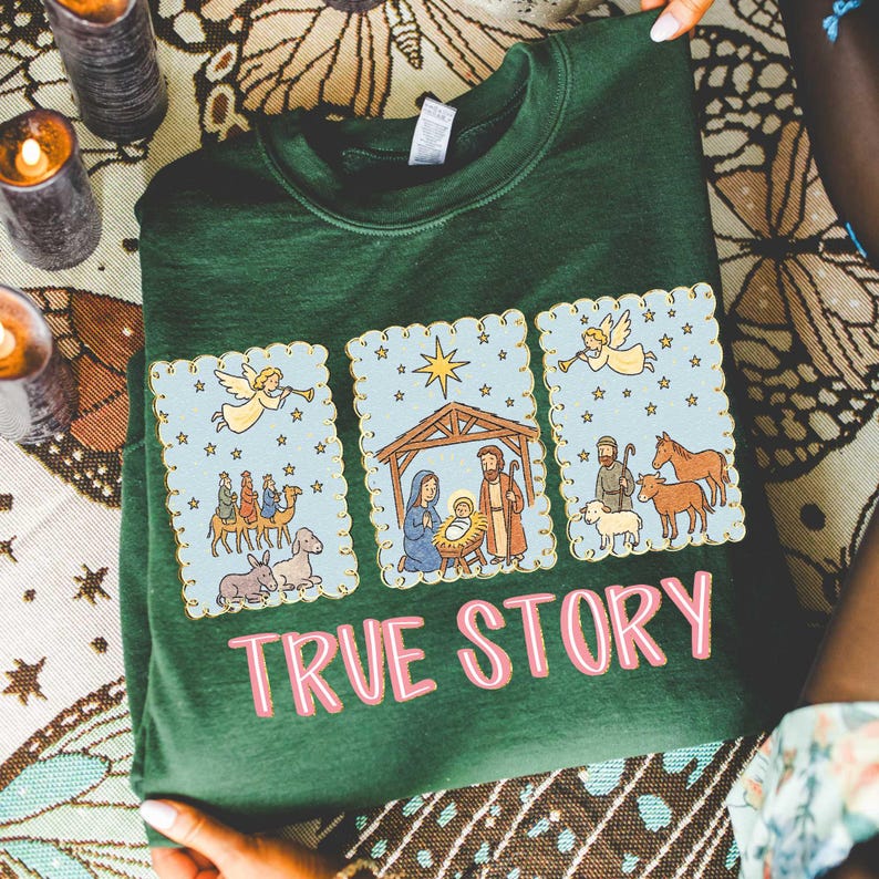 May include: A dark green sweatshirt featuring a three-panel design with a religious theme. Each panel has a light blue background with a gold scalloped border and illustrations of angels, stars, and biblical scenes. The words "TRUE STORY" are printed in pink.