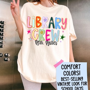 Może przedstawiać: Kremowa koszulka z napisem "LIBRARY CREW Mrs. Miller" w kolorowych, zabawnych literach. Koszulka posiada gwiazdki, kwiaty i uśmiechniętą buzię. Część kolekcji "COMFORT COLORS! BEST-SELLING VINTAGE LOOK FOR SCHOOL DAYS".