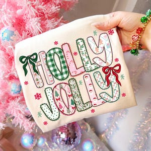 May include: A cream-colored t-shirt with the words "HOLLY JOLLY" in a festive, patterned design. The letters feature green gingham, pink, and holiday-themed patterns, with bows and snowflakes. Perfect for Christmas.