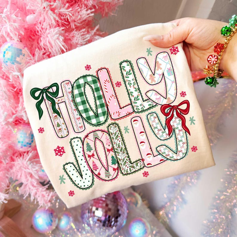 May include: A cream-colored t-shirt with the words "HOLLY JOLLY" in a festive, patterned design. The letters feature green gingham, pink, and holiday-themed patterns, with bows and snowflakes. Perfect for Christmas.
