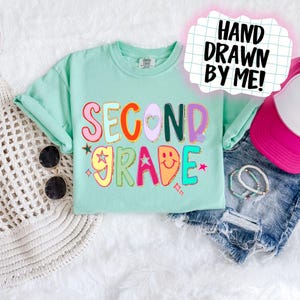 Puede incluir: Una camiseta verde menta con las palabras "SECOND GRADE" en letras coloridas y divertidas. El dise&ntilde;o incluye estrellas, corazones y una carita sonriente. El texto "HAND DRAWN BY ME!" est&aacute; en una burbuja de di&aacute;logo rosa.