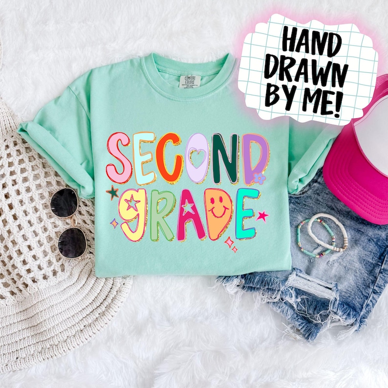 Puede incluir: Una camiseta verde menta con las palabras "SECOND GRADE" en letras coloridas y divertidas. El dise&ntilde;o incluye estrellas, corazones y una carita sonriente. El texto "HAND DRAWN BY ME!" est&aacute; en una burbuja de di&aacute;logo rosa.