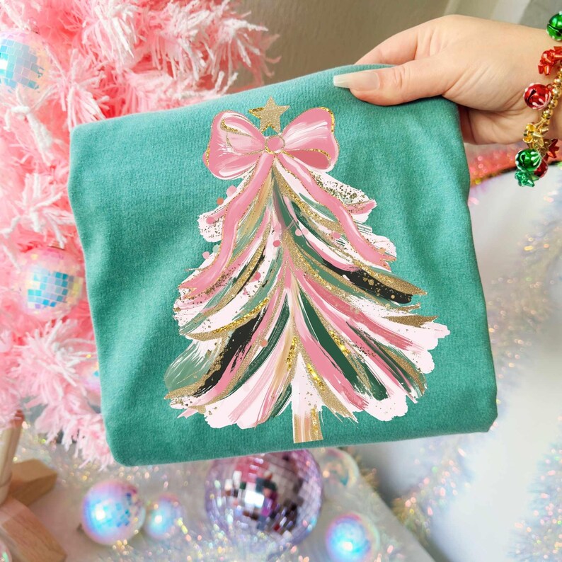 May include: Teal-colored sweatshirt featuring a Christmas tree design. The tree is decorated with pink, gold, and green brushstrokes, topped with a pink bow and gold star. The design is centered on the front of the folded garment.