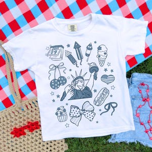Può includere: T-shirt bianco con illustrazioni patriottiche blu e grigie, tra cui la Statua della Libertà, fuochi d'artificio, gelato e un fiocco. La maglietta ha il testo "July 04". La maglietta è su una coperta a scacchi rossa, bianca e blu.
