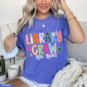 Może przedstawiać: Perwinka niebieska koszulka z napisem "LIBRARY CREW" w kolorowych, zabawnych literach. Koszulka zawiera r&oacute;wnież imię "Mrs. Miller" w białym skrypcie. Projekt zawiera gwiazdy i uśmiechniętą buzię.