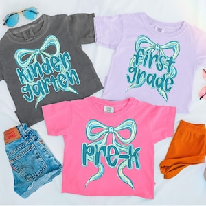 Op de afbeelding: Drie shirts met korte mouwen in grijs, lavendel en roze, elk met een groot strikdesign en schoolklas tekst. De tekst luidt "Kindergarten", "First Grade" en "Pre-K". Accessoires zijn een spijkerbroek, oranje legging, zonnebril en schoenen.