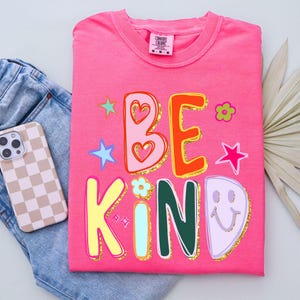 May include: A hot pink t-shirt with the words "BE KIND" in colorful, playful lettering. The design includes stars, hearts, and flowers with gold glitter accents. A smartphone with a checkered case and blue jeans are next to the shirt.