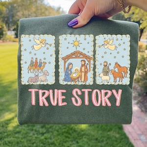 May include: Dark green t-shirt with a graphic featuring three framed illustrations of the Nativity scene. The illustrations include angels, the Three Wise Men, Mary, Joseph, and baby Jesus. The text "TRUE STORY" is printed in pink.