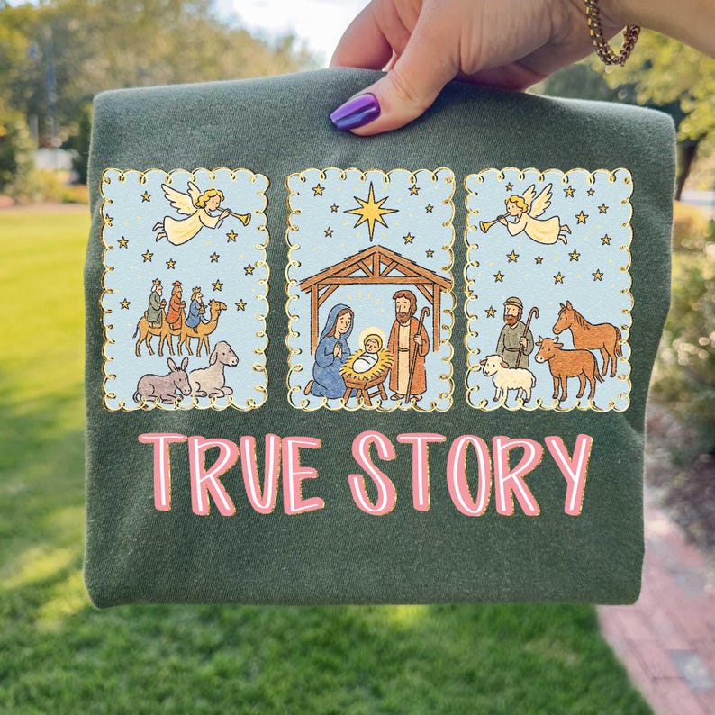May include: Dark green t-shirt with a graphic featuring three framed illustrations of the Nativity scene. The illustrations include angels, the Three Wise Men, Mary, Joseph, and baby Jesus. The text "TRUE STORY" is printed in pink.
