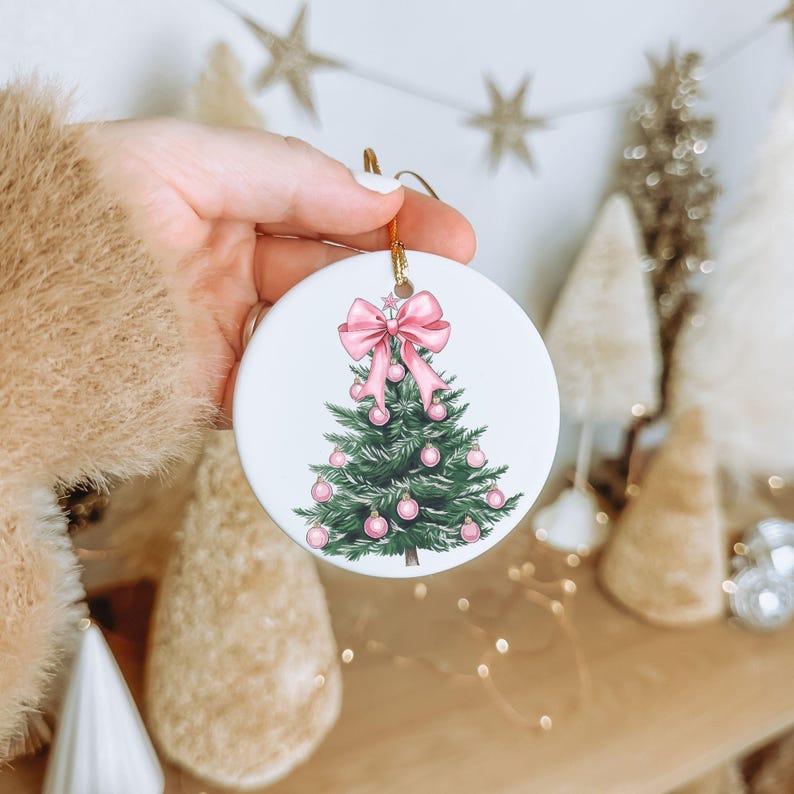 May include: A round white Christmas ornament with a gold string. The ornament features a green Christmas tree decorated with pink ornaments and a pink bow at the top. The ornament is held in a hand.