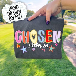 May include: Dark gray t-shirt with the word "CHOSEN" in colorful, outlined letters, accented with stars and flowers. The text "2 Peter 2:9" is below the word. A hand-drawn text bubble says "HAND DRAWN BY ME!"