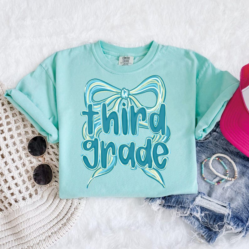 Op de afbeelding: Een lichtblauw T-shirt met de tekst "third grade" in een speels lettertype, versierd met een strik ontwerp. Het shirt maakt deel uit van een back-to-school collectie. Zonnebril, armbanden en een roze pet zijn ook zichtbaar.