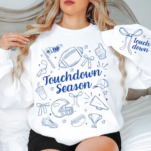 May include: White sweatshirt with blue graphic illustrations of football-related items, including a football, helmet, and trophy. The words "Touchdown Season" are prominently displayed. The sleeve features the words "touch down" with a bow.
