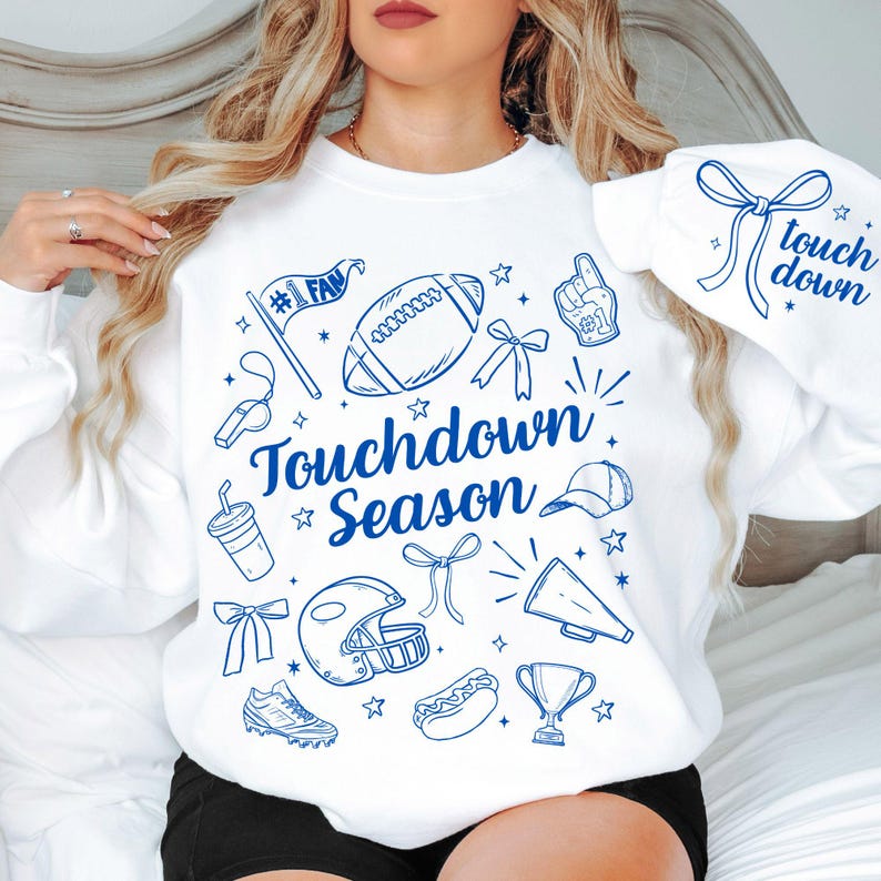 May include: White sweatshirt with blue graphic illustrations of football-related items, including a football, helmet, and trophy. The words "Touchdown Season" are prominently displayed. The sleeve features the words "touch down" with a bow.