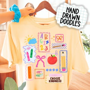Puede incluir: Una camiseta amarilla clara con garabatos coloridos dibujados a mano con temas escolares. El diseño incluye una manzana, crayones, un lápiz y las palabras "CHOOSE KINDNESS". La camiseta está colgada en una percha de madera.