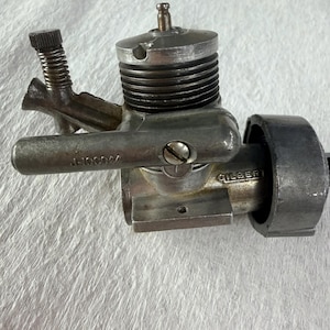 May include: A silver-toned metal model engine featuring a finned cylinder head, a threaded adjustment screw, and a side-mounted intake tube. The side of the engine body is stamped with the text GILBERT and the alphanumeric code J-100044.