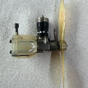 May include: A small glow plug engine for model aircraft featuring a metallic body, a black finned cylinder head, a translucent off-white plastic fuel tank section, and a long, slender, pale yellow two-blade propeller attached to the front shaft.