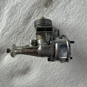 May include: A silver-colored metal internal combustion engine for a remote-controlled vehicle, featuring a finned cylinder head, a rectangular exhaust port, and the word FURY embossed on the side of the crankcase, with several mounting bolts.