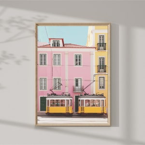 May include: Two yellow vintage trams with windows and doors are parked in front of a pink and yellow building with balconies. The building has a blue sky in the background.