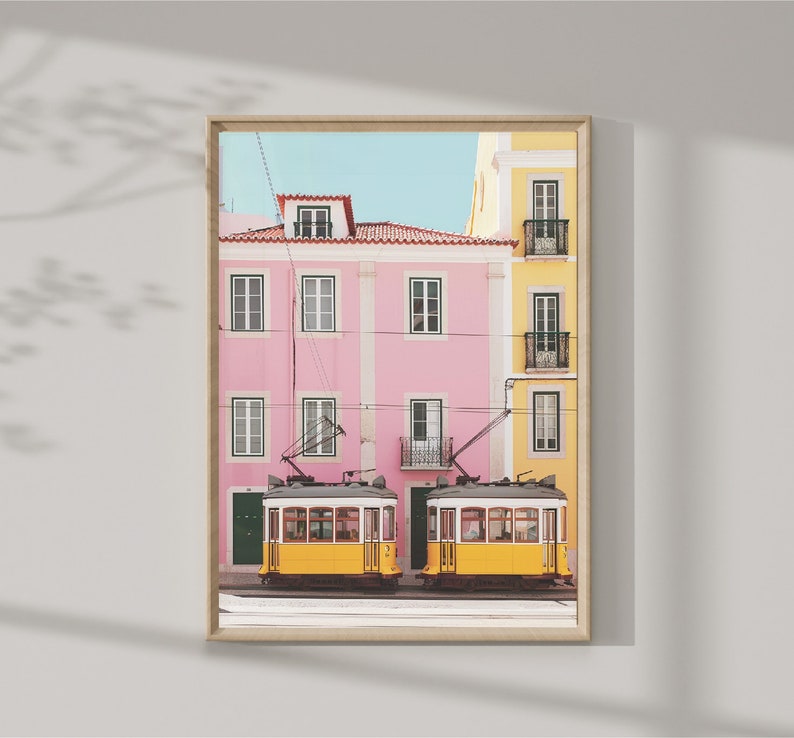 May include: Two yellow vintage trams with windows and doors are parked in front of a pink and yellow building with balconies. The building has a blue sky in the background.