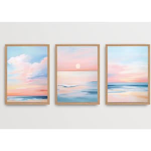 May include: A set of three framed paintings depicting a seascape with a pink and blue sunset over the ocean. The paintings are in wooden frames and are hung on a white wall.