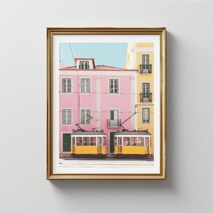 May include: Two yellow vintage-style streetcars with black trim are parked in front of a pink and yellow building. The building has a balcony and windows. The streetcars are in front of a building with a balcony and windows.