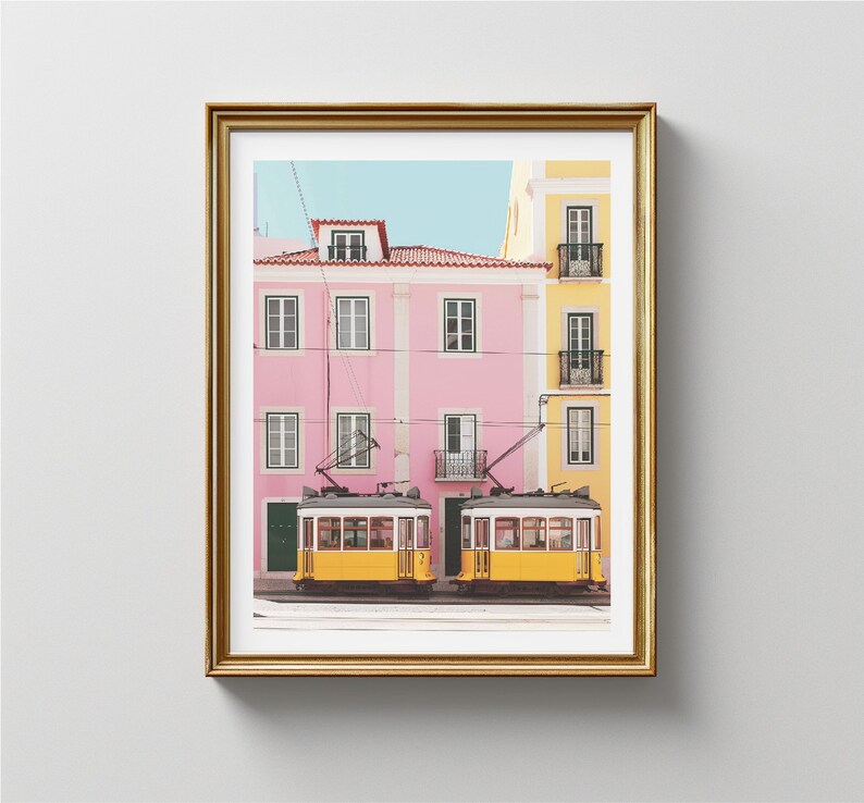 May include: Two yellow vintage-style streetcars with black trim are parked in front of a pink and yellow building. The building has a balcony and windows. The streetcars are in front of a building with a balcony and windows.