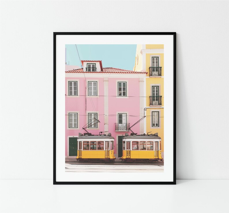 May include: Two yellow vintage streetcars parked in front of a pink and yellow building with a blue sky in the background.