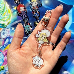 Puede incluir: Llaveros de acrílico transparente con personajes de estilo anime. Los llaveros tienen cierres metálicos plateados. Los personajes tienen varios colores de pelo y atuendos. Un llavero presenta un animal blanco y esponjoso. El fondo es una superficie estampada azul y verde.